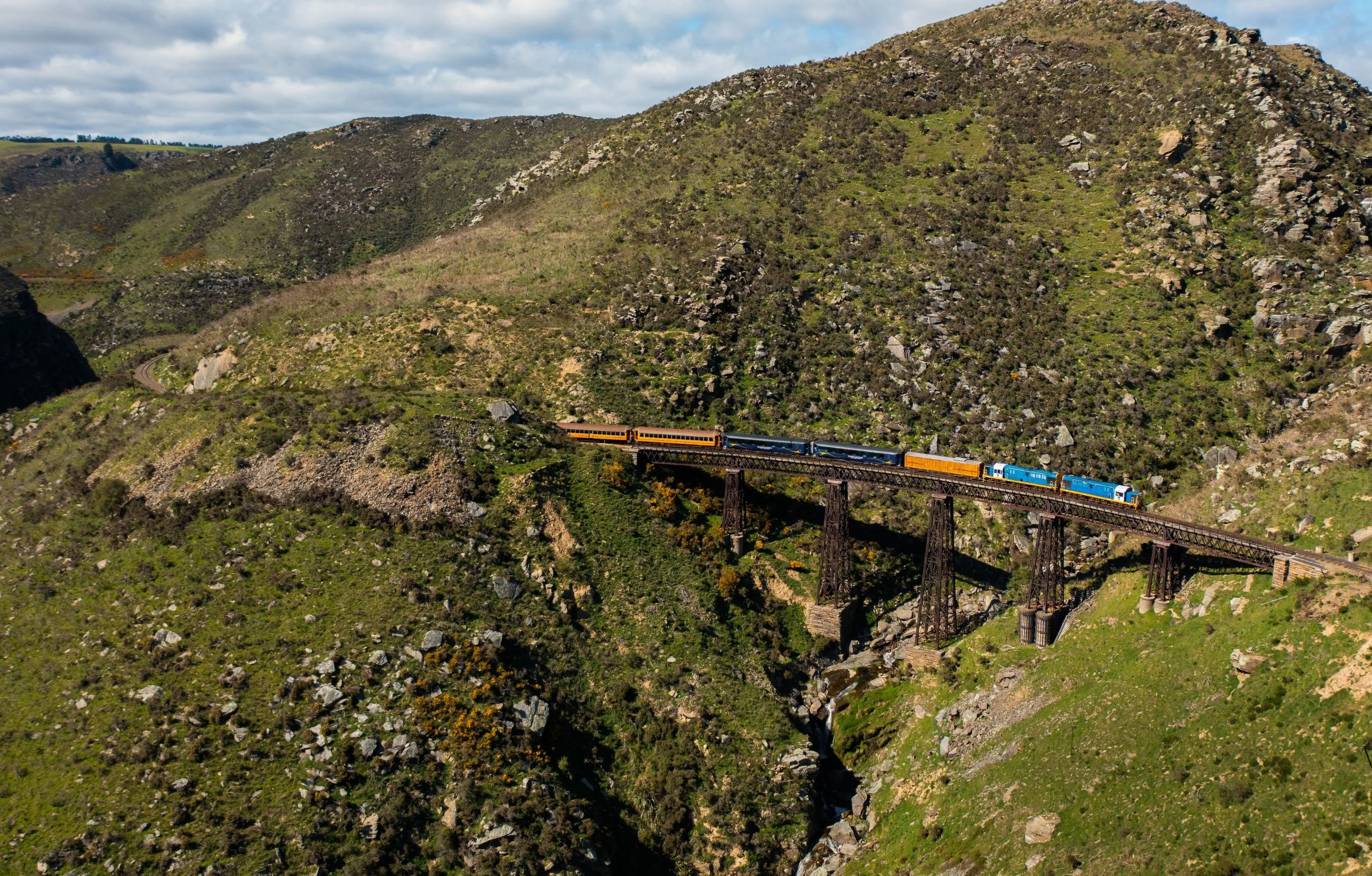A blue and yellow train crosses a tall steel viaduct spanning a rocky, grassy gorge between rolling hills.